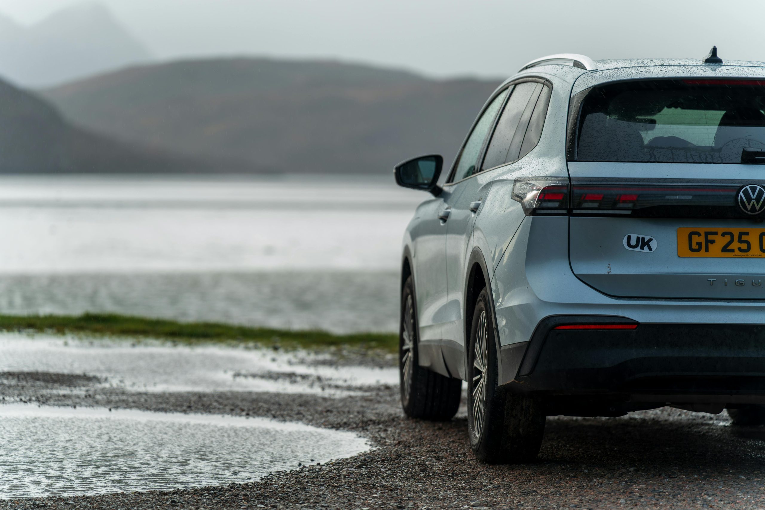 A silver SUV parked by a serene lakeside with mountainous background under a gray sky.