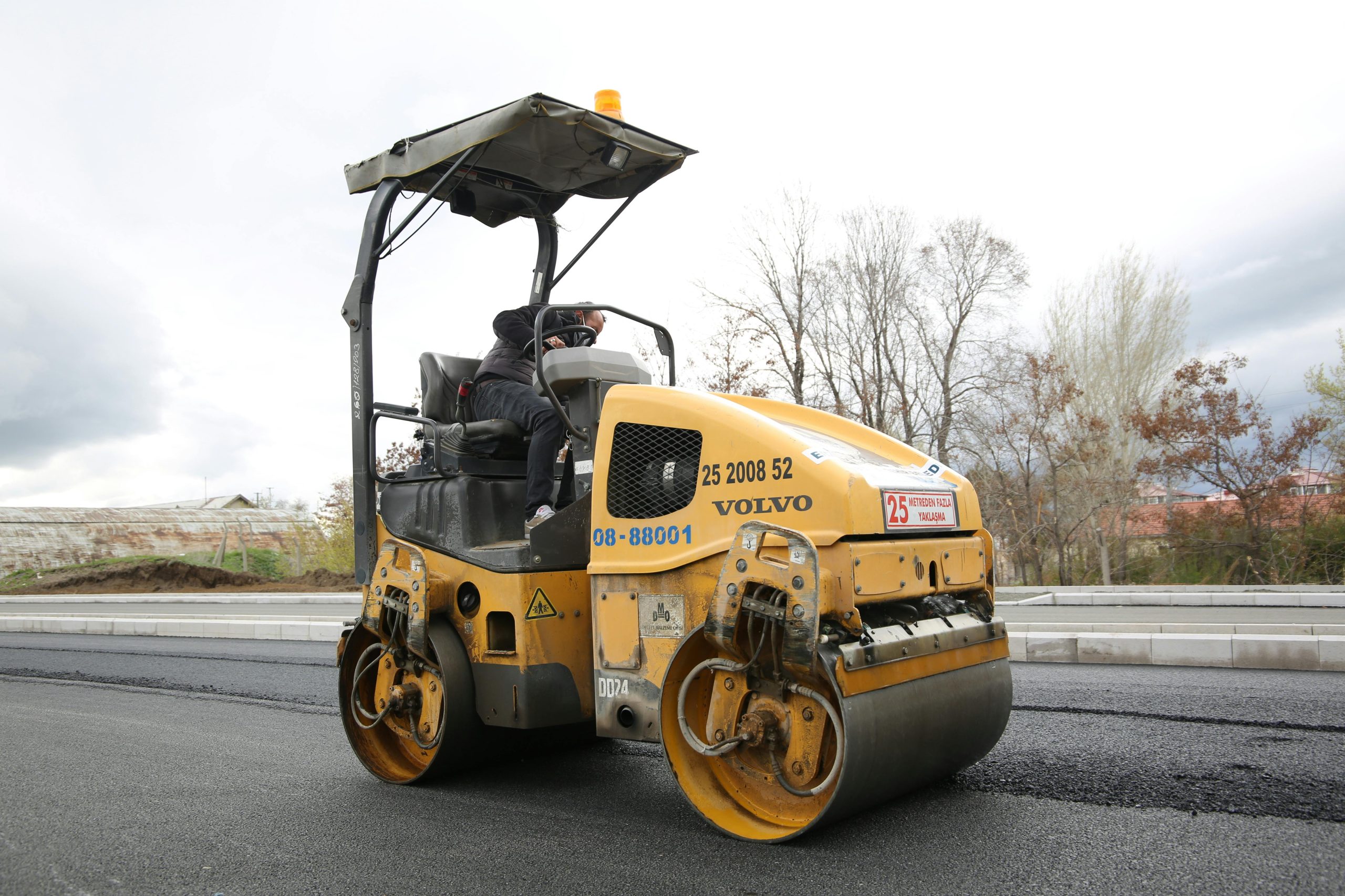 A road roller compacting fresh asphalt on a new roadway with a construction worker operating the vehicle.