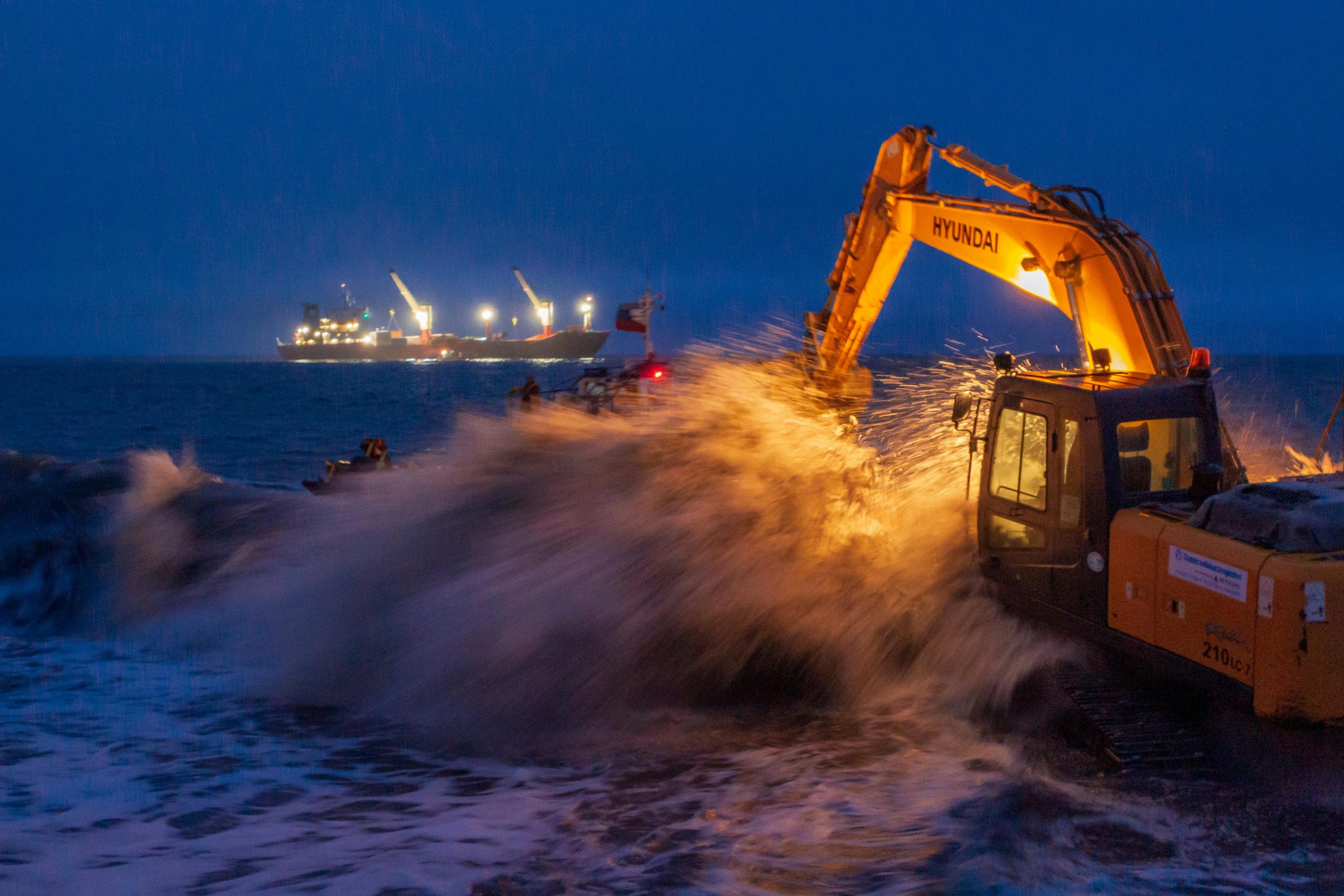 An excavator operates by the sea at night, splashing waves against its arm under vivid ship lights.